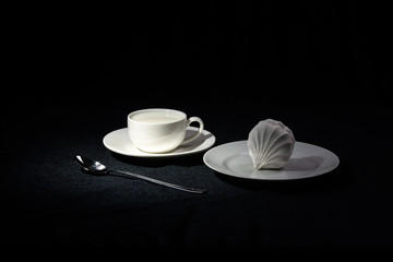 A saucer with a coffee Cup filled with milk and a plate with marshmallows on a black cloth background