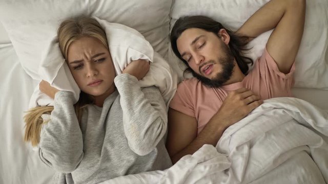 A displeased disappointed young woman is covering her ears with a pillow while her boyfriend is snoring in the bed at the bedroom