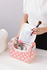 Cropped shot of a lady standing near the table with a makeup brush and open makeup bag with skin care products. The pink beauty pouch with white polka dot print is equipped with a zip fastener. 