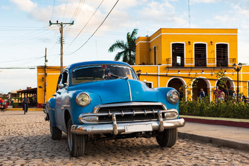 classic blue car on cobblestone road in front of colorful houses in trinidad, cuba