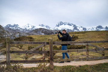 young man taking a picture from a bridge with scenic views of snowed mountains and green hills...