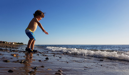 Four year old boy sitting by the sea at sunset, playing with water