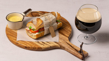 Ham, tomato, sandwich with soft cheese wrapped in a paper lying on a wooden cutting board. There is a glass of dark beer near it. 