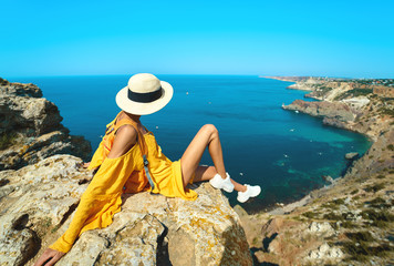 side view of traveler woman in bright yellow dress and hat sitting on cliff edge with beautiful sea view and enjoying wonderful nature