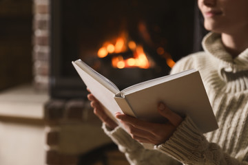 Woman reading book near burning fireplace at home, closeup