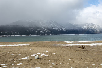Fototapeta na wymiar Sparse snow on beach with forgotten tree stump and fog shrouded mountain range on cloudy day