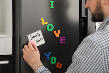 Man putting note with phrase SMILE MORE on refrigerator door, closeup