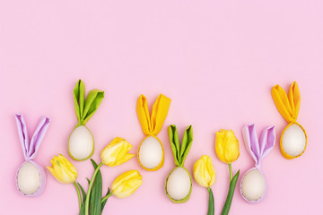Colorful Easter eggs with cloth ears as Easter bunny. Bright yellow spring flowers tulip on pink background with copy space. Holiday spring concept.