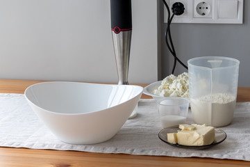 A white bowl, a hand blender, plugged in the power socket, containers with flour, butter, egg, sugar, baking powder, cottage cheese stand on a table. The process of making curd bagels or croissants
