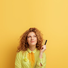 smiling redhead woman holding credit card while thinking on yellow