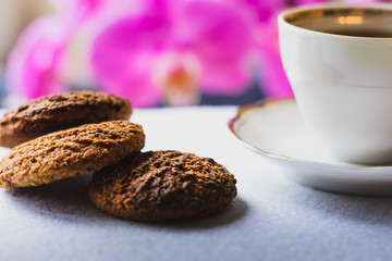oatmeal cookies with a white cup on a white fabric tablecloth, purple orchid stands in the background