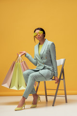 attractive african american woman in suit and sunglasses with flowers holding shopping bags while sitting on yellow