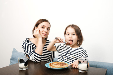 Obraz na płótnie Canvas Mother and little daughter eating pancakes with milk in the morning in light kitchen with white background