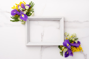Fresh purple irises and yellow flowers bouquet, blank photo frame on white wooden background on white marble background. Spring, easter concept. Flat lay, top view, copy space.