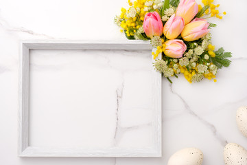 Fresh red and yellow tulips flowers bouquet, blank photo frame and eggs on white wooden background on white marble background. Spring, easter concept. Flat lay, top view, copy space.