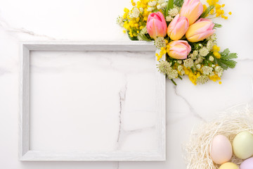 Fresh red and yellow tulips flowers bouquet, blank photo frame and eggs on white wooden background on white marble background. Spring, easter concept. Flat lay, top view, copy space.