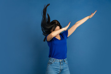 Obraz na płótnie Canvas Dabbing. Beautiful caucasian woman's portrait isolated on blue studio background. Beautiful female brunette model in casual style. Concept of human emotions, facial expression, sales, ad, copyspace.