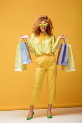 stylish redhead woman in sunglasses with flowers holding shopping bags on yellow