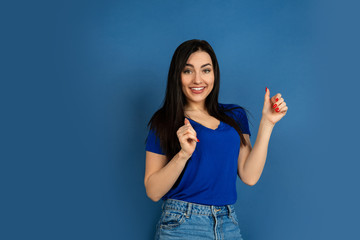 Thumb up. Beautiful caucasian woman's portrait isolated on blue studio background. Beautiful female brunette model in casual style. Concept of human emotions, facial expression, sales, ad, copyspace.