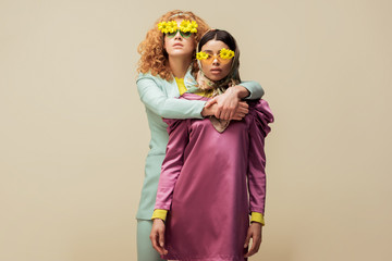 attractive redhead woman in sunglasses with flowers hugging trendy african american girl isolated on beige