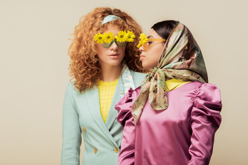 trendy african american girl and curly redhead woman in sunglasses with flowers posing isolated on beige