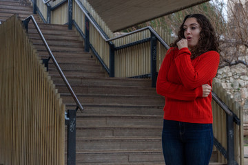 Portrait of pretty caucasian young woman holds hands in fists under chin, looks worried, has eyes popped out, being frightened by unexpected news, in the park, orange sweater and jeans, long curly hai