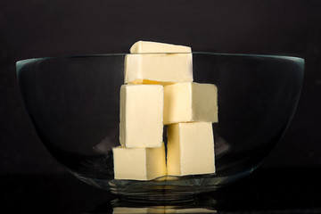 Pieces of butter lie in a glass bowl on a dark background in the studio.