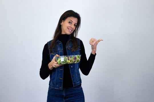 Portrait of attractive middle age woman model with salad pointing aside with cheerful expression, showing something amazing at blank space, isolated on gray background studio shot, black sweater, deni