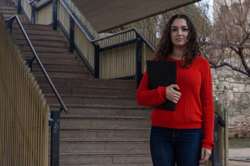 Portrait of attractive young woman model with a folder in her hands, in the park, orange sweater and jeans. Place for your text in copy space.