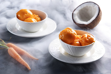 Desserts. Fruit ice cream in a white bowl next to coconut and carrots. White smoke. Background image, copy space