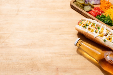 delicious hot dog with corn, green onion and mayonnaise near bottle of beer and board with vegetables on wooden table