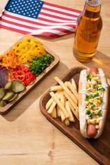 delicious hot dog near american flag, board with sliced vegetables, beer and french fries on wooden table