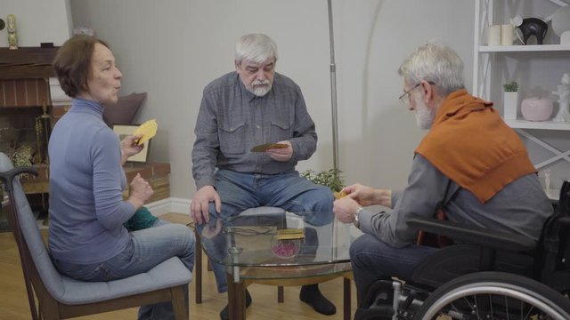 Portrait of senior Caucasian man playing cards with friends in nursing home. Retirees enjoying evening indoors. Focus changes from background to front.