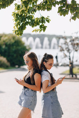 Naklejka premium two young pretty girls on a walk in the park with phones. sunny summer day, joy and friendships.