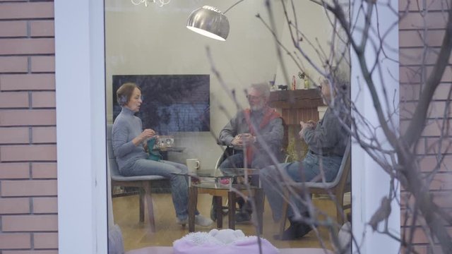 Three elderly Caucasian people chatting and drinking tea in nursing home. Men and woman spending quiet evening indoors. Wide shot, shooting from outside.
