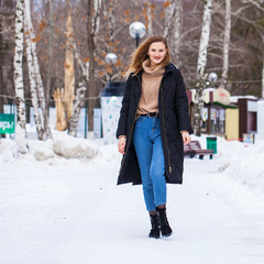 Full-length portrait of a young woman in black long down jacket posing in winter park