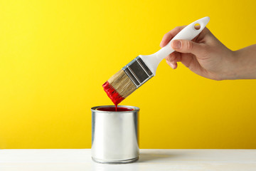 Female hand holding brush over the can with red paint against yellow background