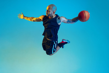 Fototapeta na wymiar In jump. Young basketball player of team wearing sportwear training, practicing in action, motion on blue background in neon light. Concept of sport, movement, energy and dynamic, healthy lifestyle.