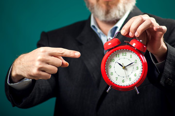 Bearded businessman holding alarm clock and pointing with a finger at it. Time management concept