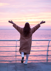 A young girl in a fur coat with a hot drink welcomes a red sunrise on the sea promenade