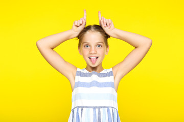 Young girl showing tongue and making rabbit ears by fingers on yellow background