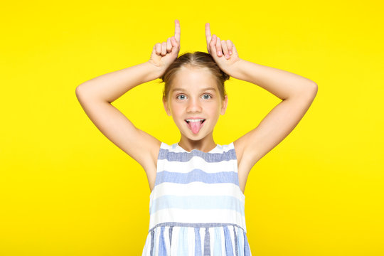 Young Girl Showing Tongue And Making Rabbit Ears By Fingers On Yellow Background