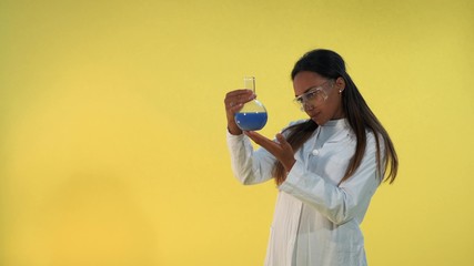 Black female scientist in lab coat looking on flask with experimental liquid on yellow background....