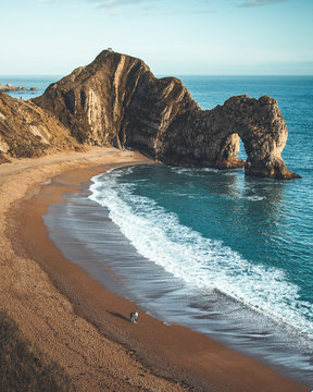 Couple Walking On The Beach With Ancient Rock Formation. Durdle Door, Dorset.  People Walking, Active On Beach. Isolated.