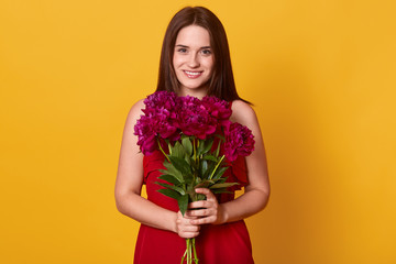 Horizontal shot of beautiful brunette woman with flower, winsome female posing with bouquet in hands, lady wearing red summer dress, adorable girl posing isolated over yellow studio background.