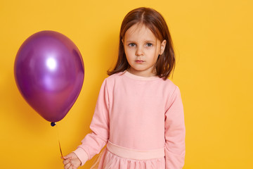 Fototapeta na wymiar Horizontal indoor portrait of cute good looking little child looking directly at camera, having holiday, standing isolated over yellow background, holding pink balloon, holding direclty at camera.