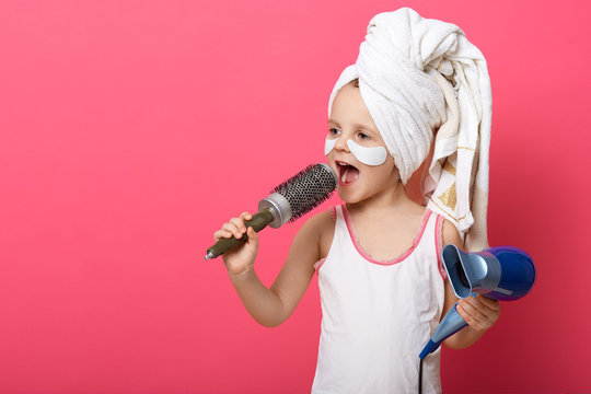 Cool little girl with hair dryer in hands. Portrait of beautiful female child singing with hair comb in hand isoalted on rosy background, charming little kid having fun in batroom after taking shower.
