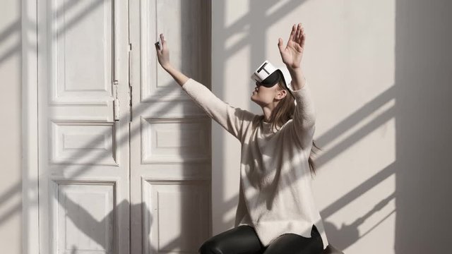 Beautiful young female playing game in vr glasses on sunny day. Happy woman touch something using modern 3D virtual reality headset on white background indoors. Augmented Reality. Future is now 4K