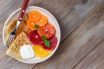Crepes  with citrus and sour cream for breakfast. Thin pancakes with slices of orange and mandarin on a plate on a wooden table. Copy space. Top view