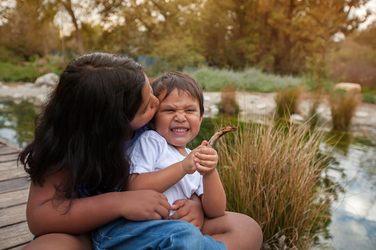 A little brother that is grimacing after his older sister gives him a kiss, both are sitting together near a pond during a sunset.